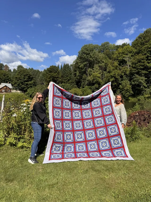 Two neighbors holding up a handmade quilt in a field by the pond.